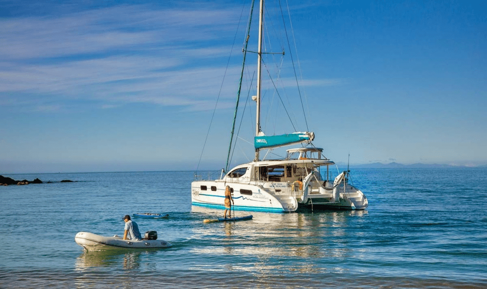 a sunny day with turquoise water and a sail boat, with a woman paddleboarding in the centre of the picture, and a man in a rubber dinghy with an outboard motor puttering towards shore at the edge of the frame