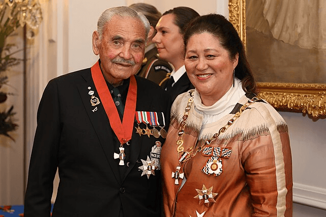 An elderly man and woman are standing together, both wearing formal attire adorned with various medals and decorations. The setting appears to be a formal event or ceremony with other people in the background.