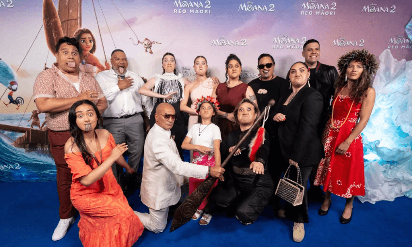 A group of people dressed in traditional Maori attire pose playfully on a blue carpet for a "Moana 2 Reo Māori" event. The backdrop features animated scenes from the movie. Some individuals make expressive faces and gestures.