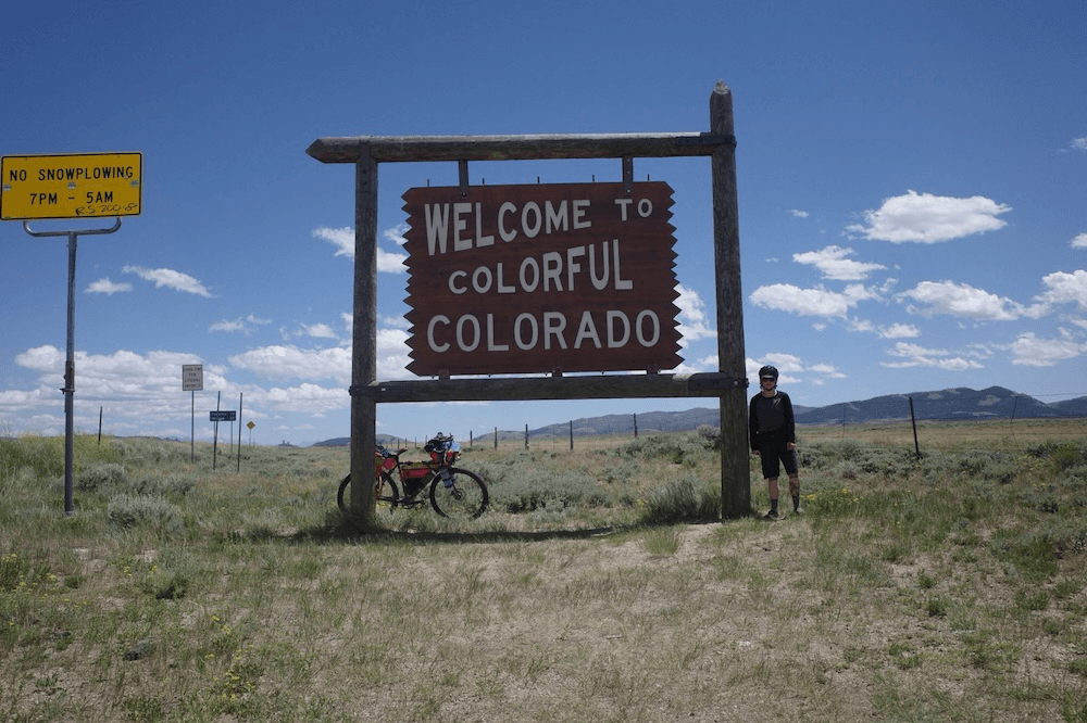 Robbie wheb, a man in a black shirt, looks small and backlit beside a sign saying 'welcome to colorful colorado', with his bike leaning against a post, and lots of bluesky and the start of some mountains in the background