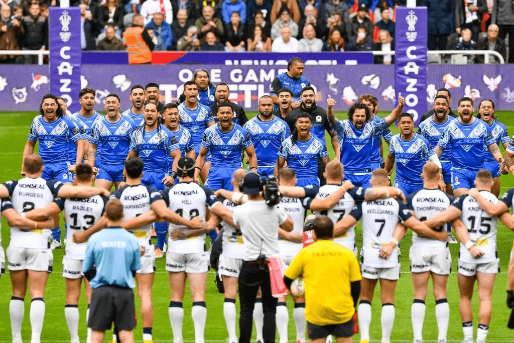 A rugby league team in blue performs a siva tau on the field, facing an opposing team in white. The crowd watches in the background during a match.