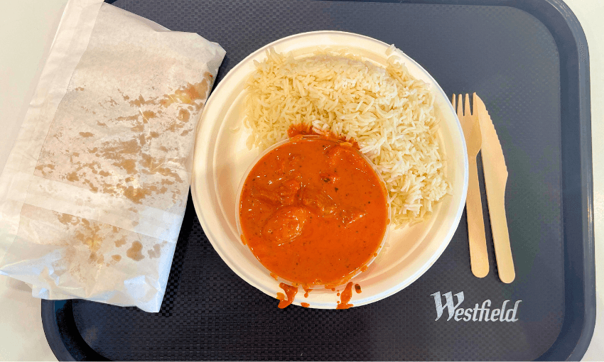 A plate of rice and orange butter chicken curry sits on a tray with a wrapped naan bread on the side. Wooden fork and knife are placed on the tray, which has a "Westfield" logo.