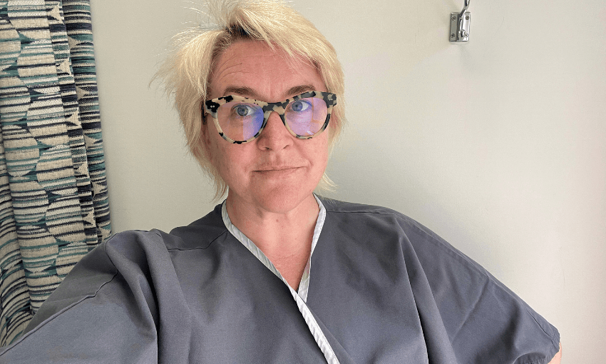 A person with short, blonde hair wearing glasses and a gray medical gown poses indoors. A patterned curtain is visible in the background.