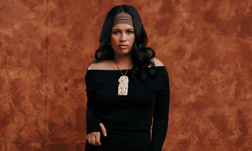 A woman with long, dark hair wears a black off-the-shoulder top and a headband. She has a large pendant necklace. The background is a textured brown backdrop.