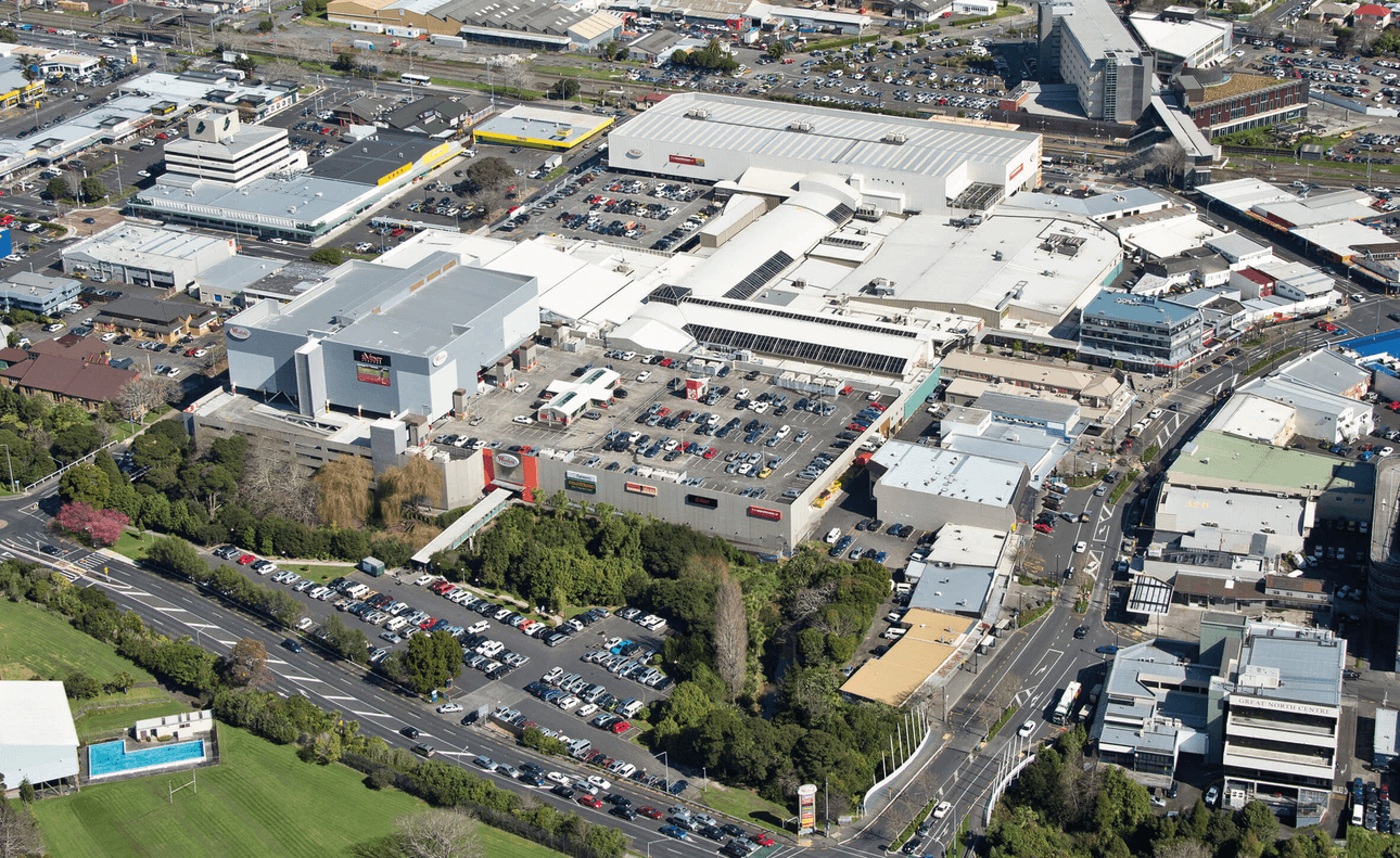 Aerial view of a large shopping mall surrounded by parking lots and roads. Numerous cars are parked nearby, with industrial and residential buildings in the background. Green spaces and trees are visible, dividing the urban landscape.