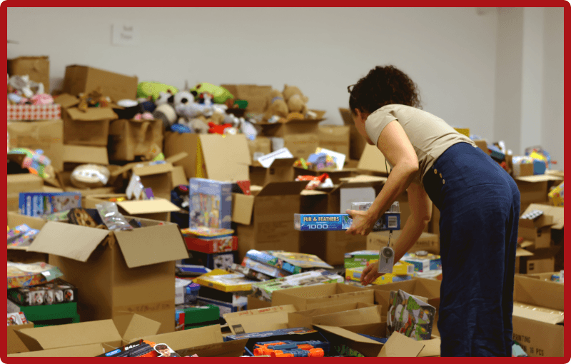 A volunteer in a beige top, dark blue jeans, selects a few toys from an area full of carboard boxes full of books, toys, games and other holiday gifts.