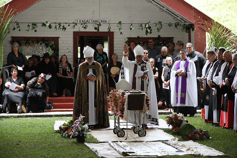 A group of clergy in ceremonial robes conducts a service outside a building adorned with greenery. A crowd of people is gathered, with floral arrangements placed around a central point. A coffin covered by a feather cloak is featured in the middle.