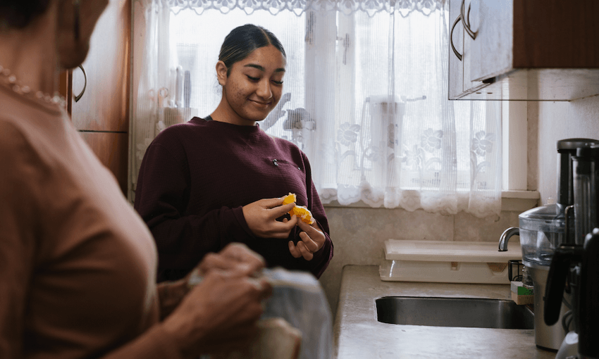 A young woman wearing a burgundy coloured jumper peels a mandarin orange. She is standing in a cream coloured kitchen with soft light streaming in behind her.