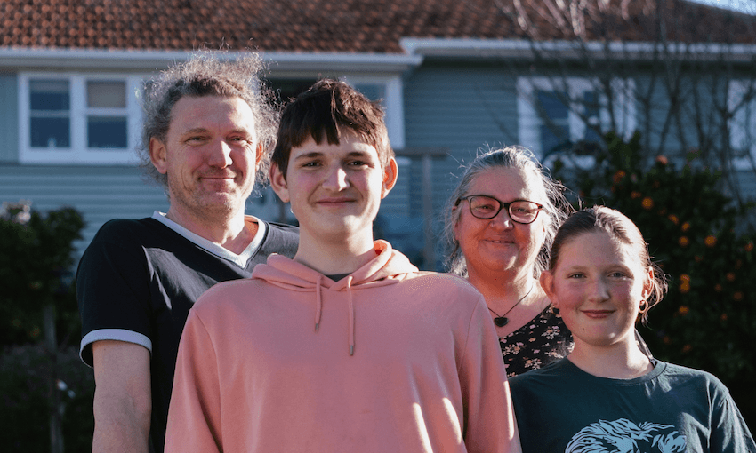 A family of four stand outside their light green home with a red roof. The son and daughter in the foreground are bothy smiling and wearing tee shirts. The mother and father stand behind.