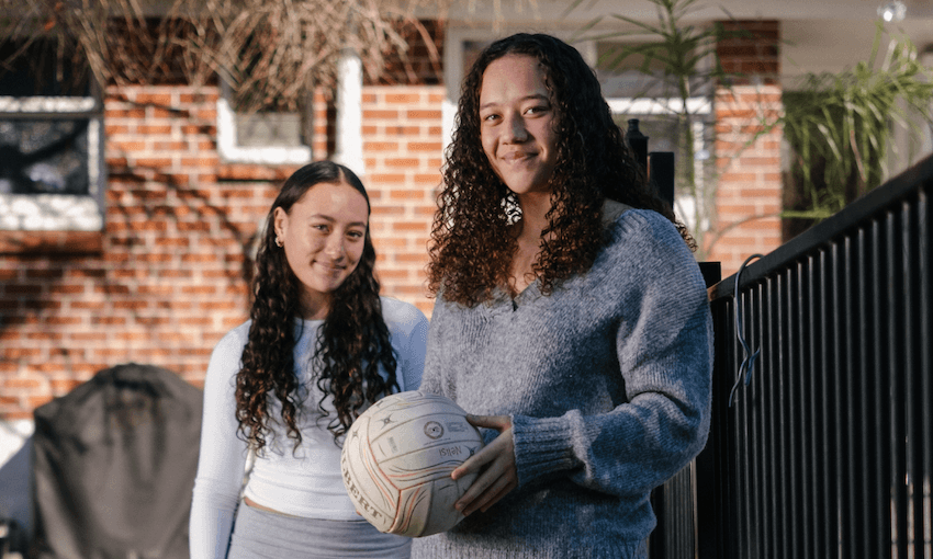 Two girls stand outside their red brick home, they have a volleyball wth them, both are smiling, have brown curly hair worn down and are wearing white tops. The are both smiling.