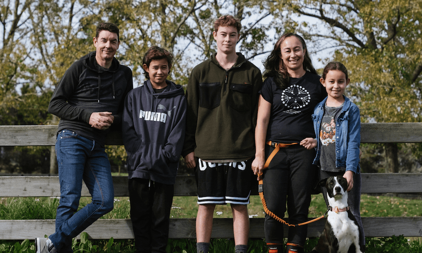 A family of five lean against a fence. They are all wearing dark coloured athletic tops and either sweatshirts or tees. Their black and white dog is with them.