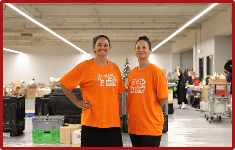 Two women, Tara Moala at left and Anataia Fu at right, wear orange Auckland City Mission tshirts and black pants and are standing side by side smiling in front of the gift and food packing stations in a large open warehouse space.