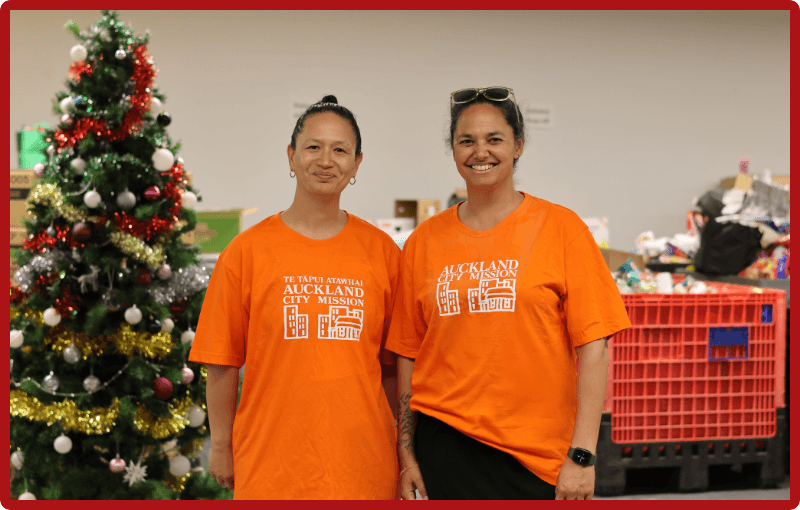 Anataia Fu and Tara Moala stand side by side in orange Auckland City Mission t-shirts. They are standing in front of a Christmas tree with tinsel and many white and red baubles. They are both smiling and there are boxes behind them which are waiting to be packed full of gifts to share with families.