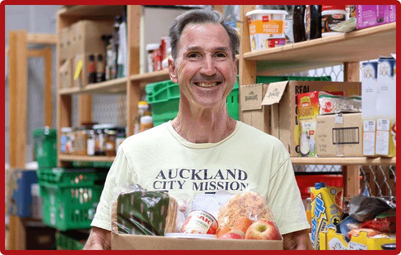 A man with gray hair in a light yellow vintage t-shirt that says Auckland City Mission on the front in black text is holding a carboard box that is filled to the brim with apples, canned goods and packets of food. Behind him are wooden shelves with food items on then. He is smiling gently