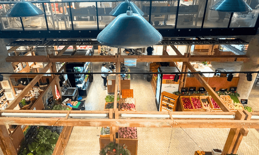 Aerial view of an indoor market with wooden beams. Various fruits and vegetables are displayed on shelves, and shoppers are browsing the produce. Large industrial lamps hang from the ceiling, casting light on the market area.