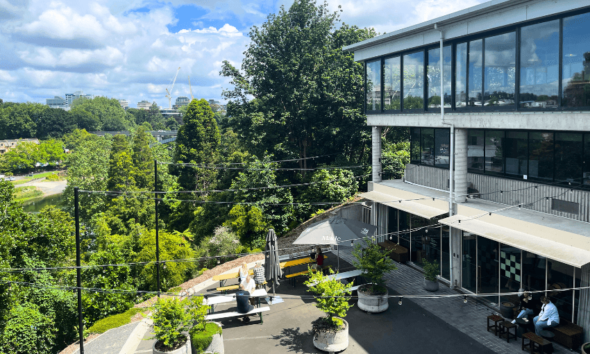 A modern building with large glass windows overlooks a lush green area. People are sitting at outdoor tables on a paved terrace. The sky is partly cloudy, and there is a river visible in the background.