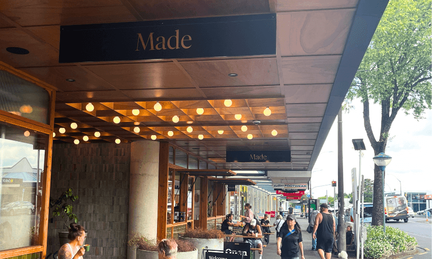 A bustling street scene outside a cafe named "Made," featuring people walking and seated under a wooden awning with hanging lights. Trees line the street, and shops are visible in the background.