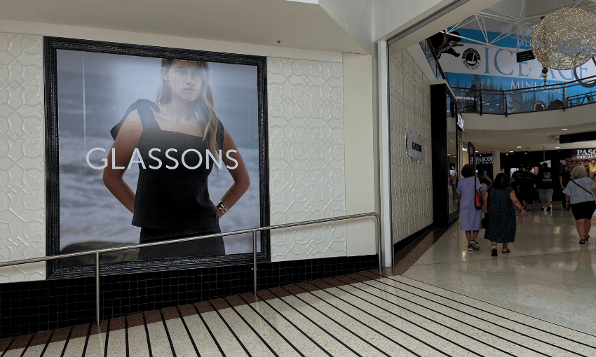 A Glassons clothing advertisement featuring a woman in a dark outfit is displayed on a mall wall. Shoppers walk by on a tiled floor, with various stores visible in the background.