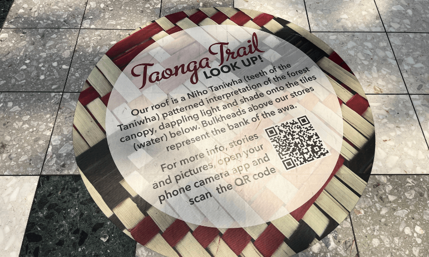 Circular sign on a patterned tile floor. It reads "Taonga Trail. Look Up!" with a description of the design as a Niho Taniwha pattern and includes a QR code for more information. The sign features red and cream colors.