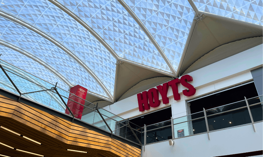 The image shows the interior of a shopping center with a domed, geometric-patterned glass ceiling. A red sign with the word "HOYTS" is visible on a wall, indicating a cinema. The architecture features wood and modern design elements.