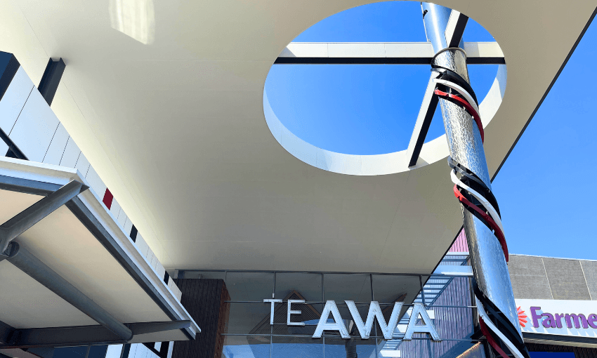 The image shows the exterior entrance of a shopping center called Te Awa. A circular opening in the roof allows sunlight to shine through. A tall, decorated pole stands on the right, and parts of other buildings are visible in the background.