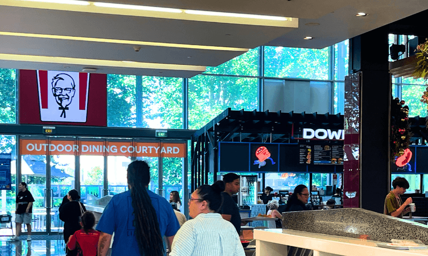 People walking inside a modern food court with glass walls. A sign indicates an outdoor dining courtyard, and a KFC logo is displayed on a screen. Bright natural light filters through the windows, highlighting the vibrant atmosphere.