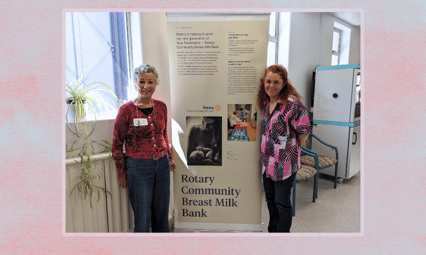 Two middle aged women, one with short curly grey hair and a red top and one with onger auburn hair and a coloured pink top, stand next to a poster reading 'rotary community breast milk bank' 