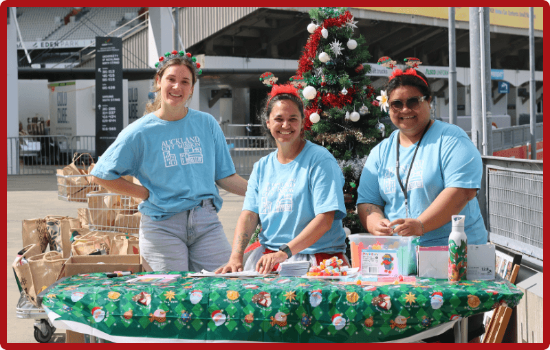 Three people stand at a table with a Christmas tablecloth on it, they are wearing baby blue Auckland City Mission t shirts and sparkly reindeer antler headbands too. There is a Christmas tree behind then and many boxes of food too to be shared with the community.