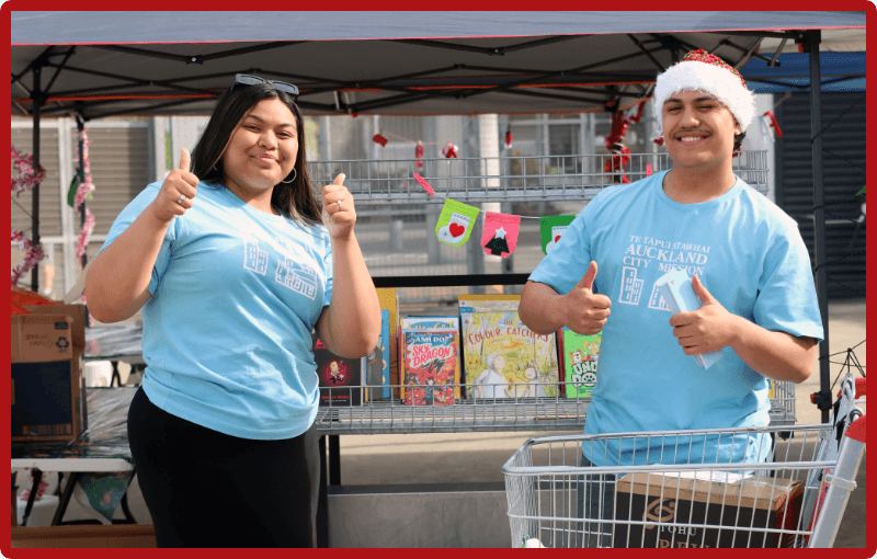 Two volunteers wearing baby blue Auckland City Mission t-shirts and santa hats are standing behind shopping carts and giving thumbs up to the camera, they are about to pack up some food boxes.