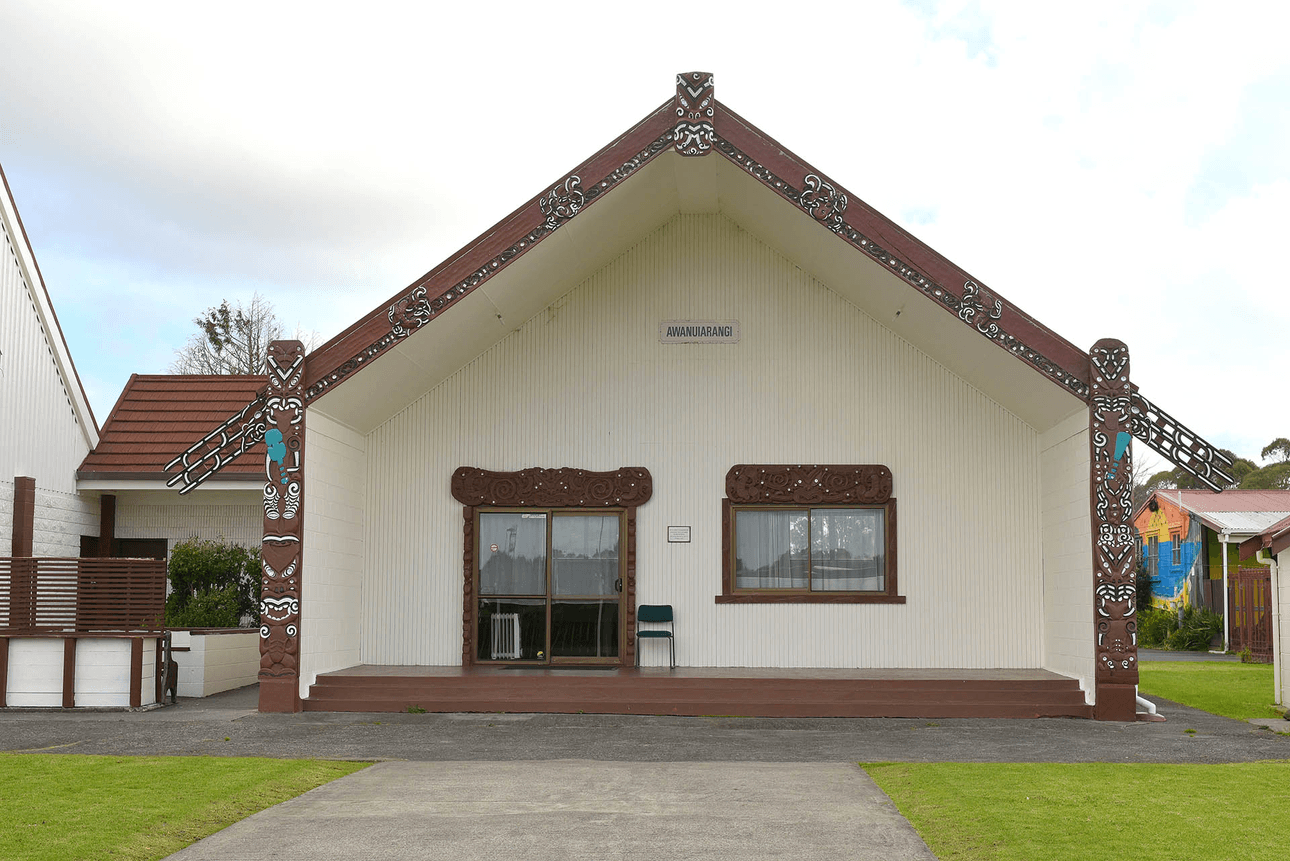 A traditional building with decorative wooden carvings on its facade, featuring a peaked roof. The structure displays intricate patterns typical of Maori architecture. Several windows and a pathway lead to the entrance.