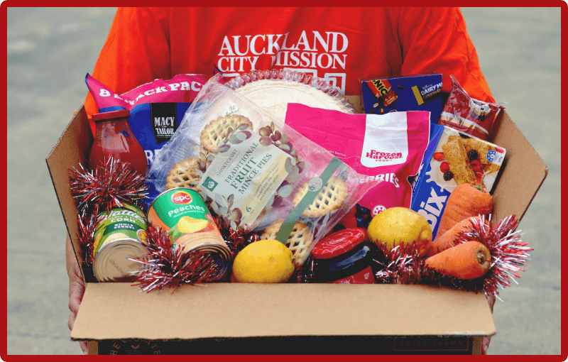 Someone wearing a red Auckland City Mission t-shirt holds a cardboard box that is overflowing with Christmas kai: minced pies in a plastic packet, canned peaches, carrots, sparkly tinsel, potatoes and more.