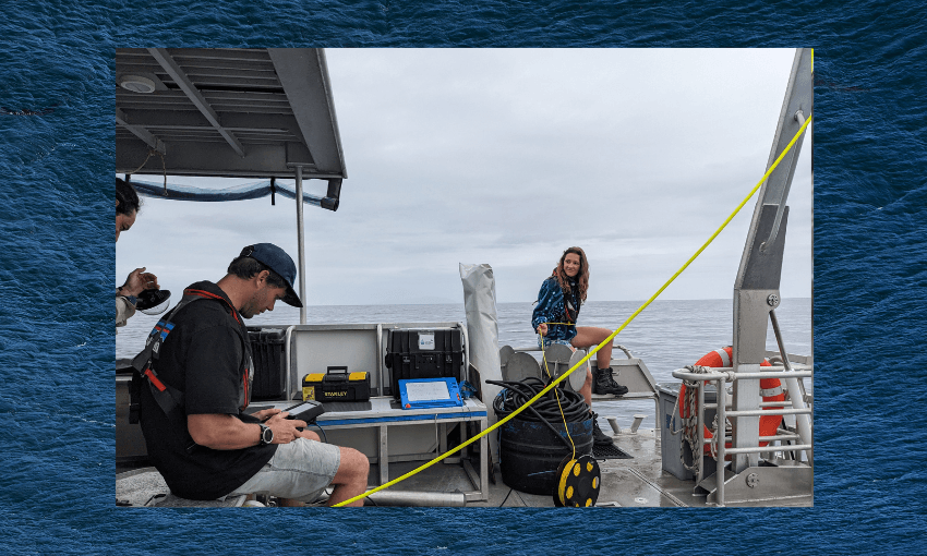 a woman in blue shorts sits at the edge of the boat looking out over the edge of the water and guiding a yellow cable, while a man with two day stubble holds a monitor 