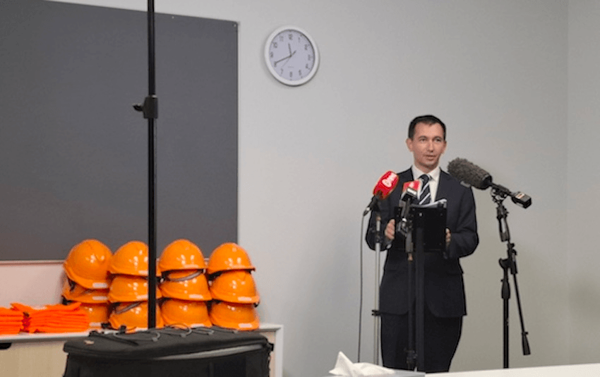 Simeon Brown wears a suit and stands in front of several microphones. Above him is a clock, to his left a pile of orange hard hats