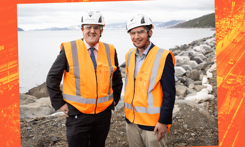 Two men wearing safety helmets and orange high-visibility vests stand on a rocky shore. The background features a body of water and a distant coastline under a cloudy sky. Both are smiling, and one has a tie.