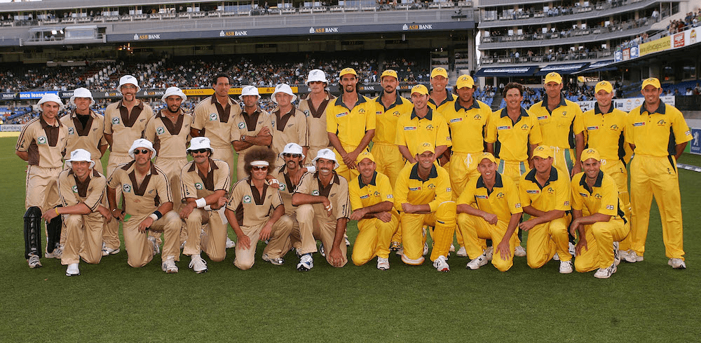 group photo of the black caps and australia teams before the first ever t20 international. nz in beige uniforms and white bucket hats, australia in yellow uniforms. several players who don't normally have a moustache have a moustache