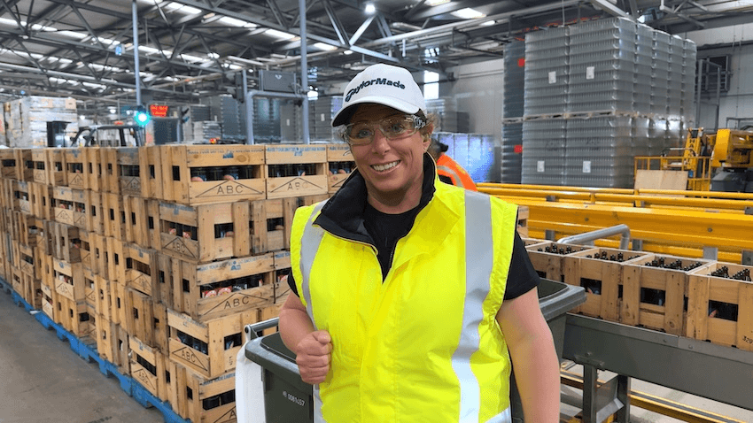 a fun and down to earth looking woman with short hair and a big smile and hi vis fest wears a cap inside a waste processing facility of some kind, with lots of cartons behind her