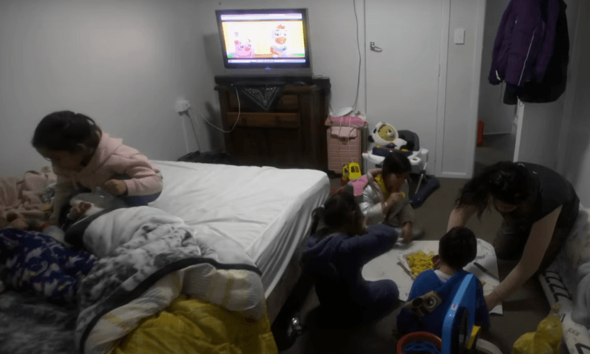 A small room with children gathered on the floor around a pile of toys and food. A woman sits with them. A bed with blankets is to the left, and a TV showing a cartoon is on a dresser. The room is cluttered with various items.