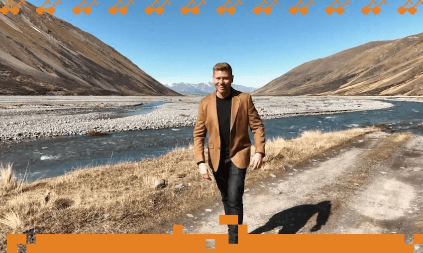 A man named Matt Gibb is wearing a brown blazer stands in front of a river in Central Otago countryside with blue skies overhead