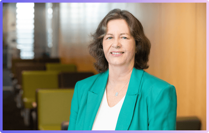 A professional portrait of Susan Watson, smiling indoors, wearing a green blazer over a white top, with a blurred background of a modern office or lounge.