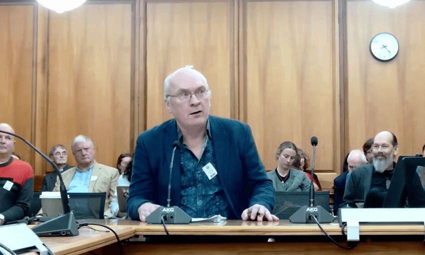 Vincent O'Malley sits at the select committee table in a suit with no tie. A small group of spectators sit behind him as he speaks into a microphone