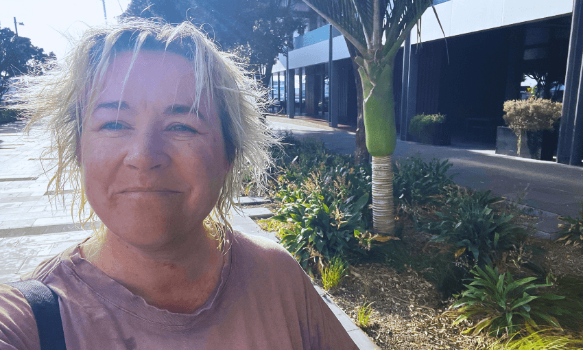 A woman (the writer) with short, light hair smiles while standing outdoors. Sunlight beams from above. They wear a casual shirt. Behind them is a palm tree and other greenery, with modern buildings in the background.