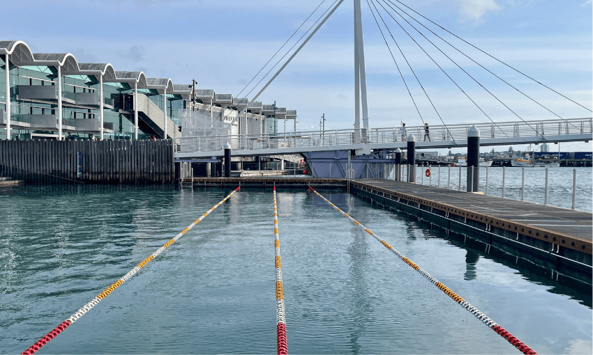 An outdoor swimming area with lane dividers is set in a harbor, positioned adjacent to a modern building and a white pedestrian bridge. The water is calm under a lightly clouded sky.