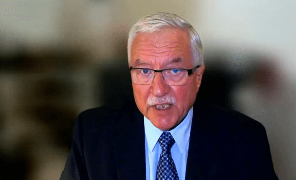 A man is centre screen on a zoom call, with white hair and white moustache and wearing a suit