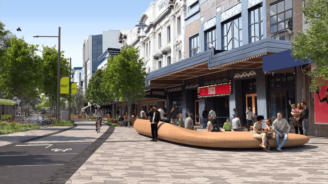 People are enjoying a sunny day in an urban plaza with modern architecture. There are trees lining the street, a bicycle lane, and wooden benches where some are sitting and talking. The area is bustling with activity and features a mix of shops and cafes.