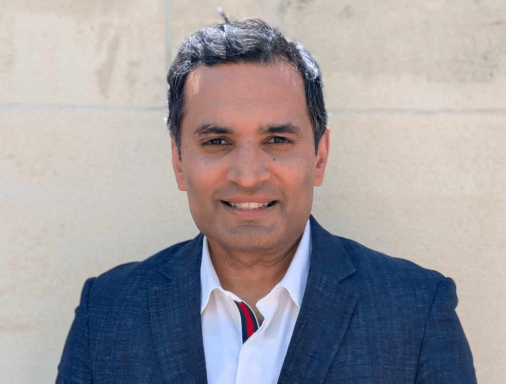 a brown man with salt and pepper hair, blazer and suit, and a slightly serious but still friendly expression