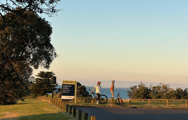 Two people with bicycles stand near a signpost in a park overlooking the ocean. The sun casts a warm glow on the scene. Trees and a clear sky are visible in the background.