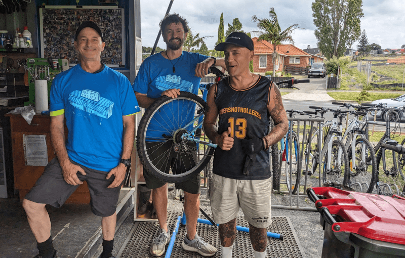 Three men smiling at a bike shop. Two wear blue t-shirts; one holds a bicycle wheel. The third is in a black sleeveless shirt with "13" on it, giving thumbs up. Bicycles and a street with houses and greenery are visible in the background.