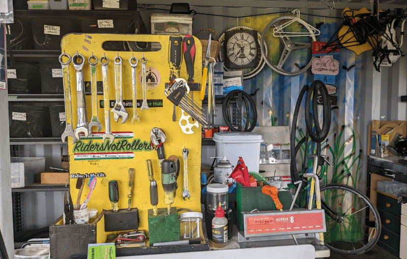 A cluttered workshop with a yellow pegboard displaying various tools like wrenches and pliers. A bicycle is mounted on the wall, surrounded by tires, boxes, and spray paint cans. A colorful, nature-themed mural covers part of the wall.