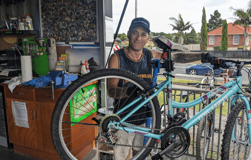 Man in a cap repairs a bicycle on a stand outside a shop. The shop has tools and supplies visible on the shelves. Cars and a house are in the background. He is smiling and wearing a dark tank top.