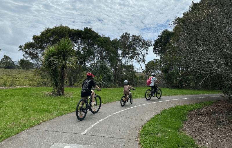 Three people of various ages ride bicycles on a paved path through a park. They are wearing helmets and surrounded by green trees and grass under a cloudy sky.